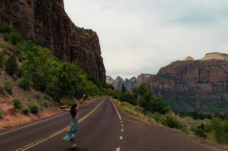 May include: A person standing on an asphalt road with arms raised, wearing a light-colored top and a green patterned skirt, set against a backdrop of towering red rock canyon walls, green trees, and a cloudy sky in a vast desert landscape.