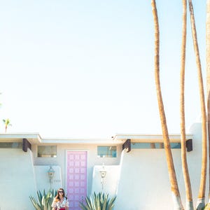 Puede incluir: Una casa blanca de estilo moderno de mediados de siglo con una puerta de entrada rosa pastel con patr&oacute;n de cuadr&iacute;cula. El exterior incluye dos grandes plantas de agave a los lados de la entrada, palmeras altas a la derecha y un cielo azul despejado.