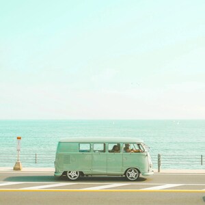 Pu&ograve; includere: Un furgone vintage color verde menta chiaro che percorre una strada costiera. Il veicolo presenta una silhouette classica e squadrata, ampi finestrini e ruote argentate. L'oceano &egrave; visibile sullo sfondo dietro un guardrail metallico.