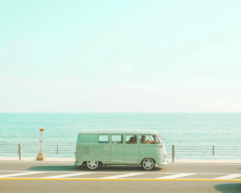 Pu&ograve; includere: Un furgone vintage color verde menta chiaro che percorre una strada costiera. Il veicolo presenta una silhouette classica e squadrata, ampi finestrini e ruote argentate. L'oceano &egrave; visibile sullo sfondo dietro un guardrail metallico.