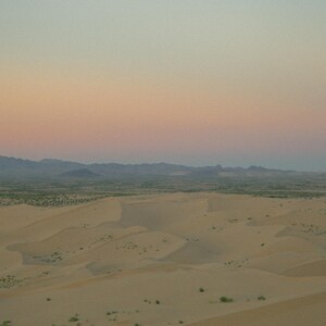 K&ouml;nnte beinhalten: Panoramablick auf eine W&uuml;stenlandschaft mit sanften Sandd&uuml;nen und einer fernen Bergkette. Der Himmel ist in sanftem Rosa und Blau gehalten und erzeugt eine ruhige und friedliche Atmosph&auml;re.