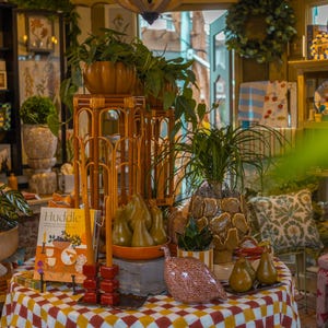 Pu&ograve; includere: Un tavolo rotondo coperto da una tovaglia a quadri rossi, gialli e bianchi presenta un libro intitolato Huddle, due alte candele rosse, una faraona in ceramica, diverse pere decorative e varie piante in vaso su supporti in bamb&ugrave;.