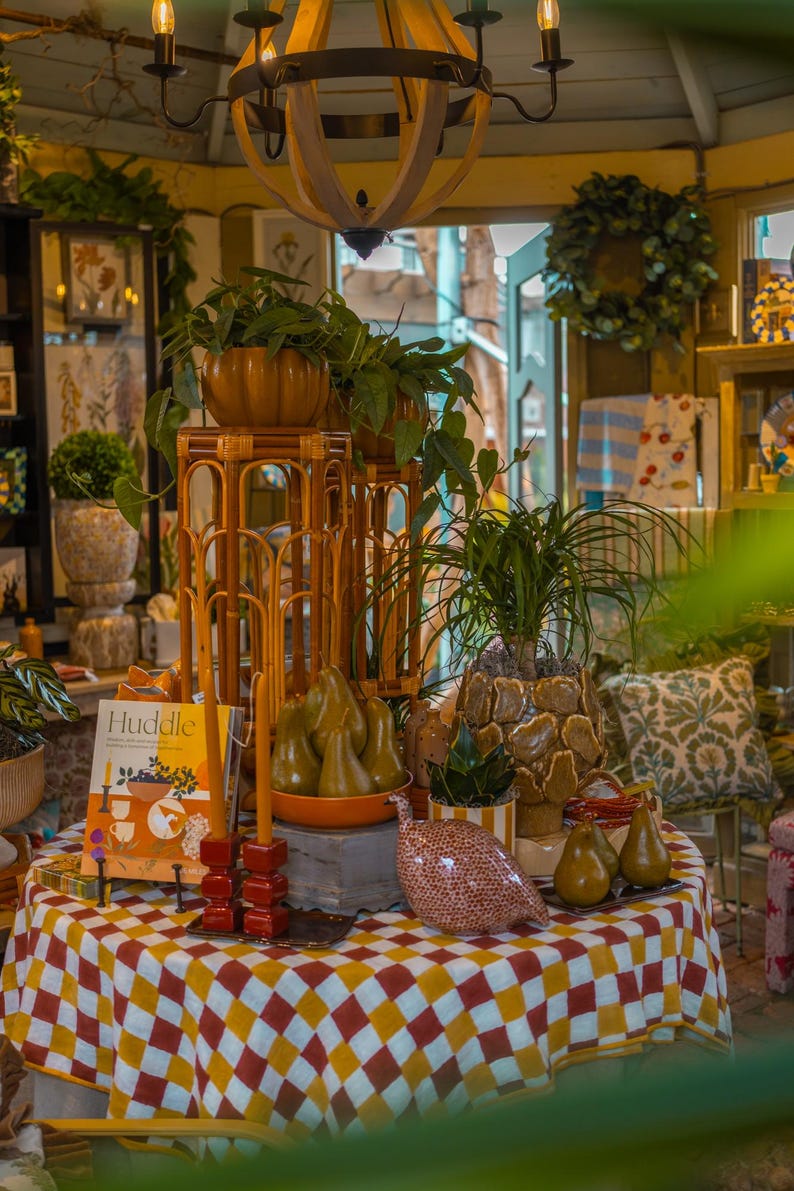 Pu&ograve; includere: Un tavolo rotondo coperto da una tovaglia a quadri rossi, gialli e bianchi presenta un libro intitolato Huddle, due alte candele rosse, una faraona in ceramica, diverse pere decorative e varie piante in vaso su supporti in bamb&ugrave;.