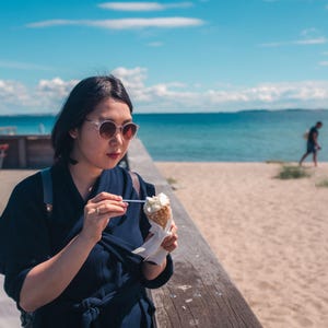 Può includere: Una persona con capelli scuri indossa occhiali da sole rotondi e un top blu scuro a portafoglio, tenendo in mano un cono gelato bianco con un cucchiaino di plastica. Il gelato è servito in una cialda croccante e avvolto in un tovagliolo di carta.