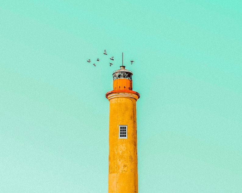 Pu&ograve; includere: Un alto faro cilindrico con un esterno giallo usurato e una sezione superiore arancione brillante. Una singola piccola finestra rettangolare &egrave; visibile sulla torre principale. Uno stormo di uccelli vola vicino alla cima contro un cielo verde menta.