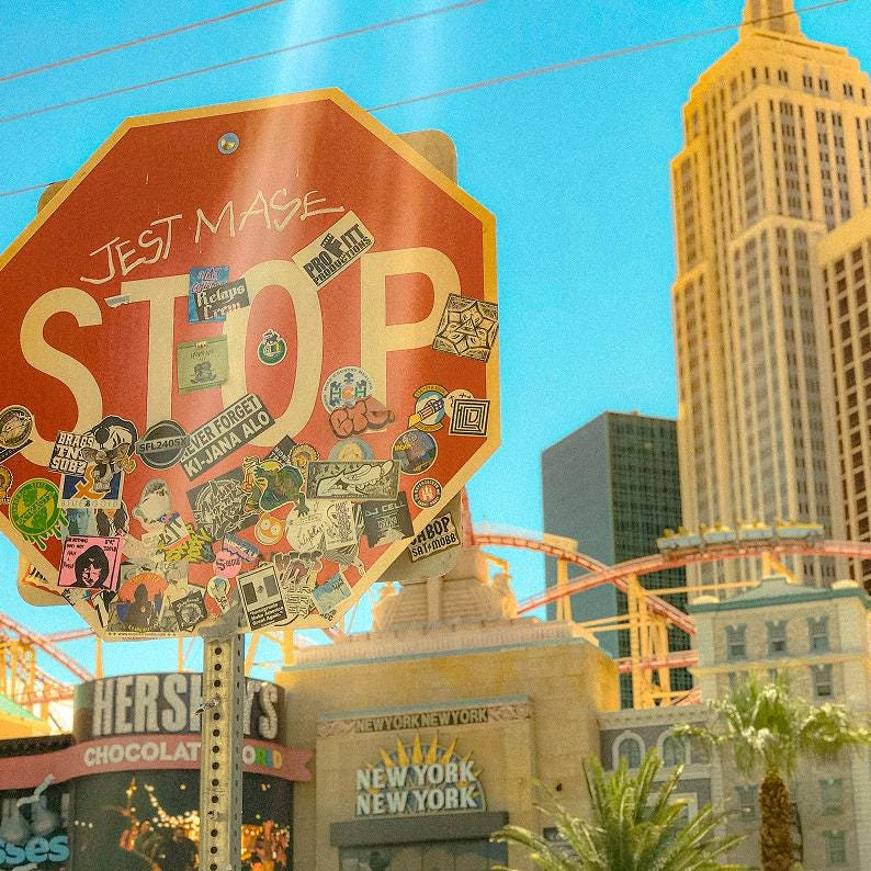 May include: A red octagonal stop sign covered in numerous colorful stickers and the handwritten text JEST MASE, positioned in front of a tall yellow skyscraper, a roller coaster track, and a building with a New York New York sign.