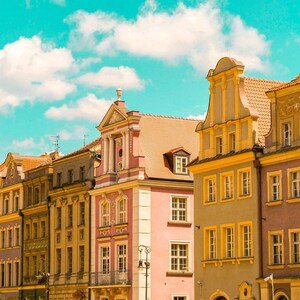 Pu&ograve; includere: Una fila di edifici a pi&ugrave; piani con facciate ornate nei toni del rosa, giallo e grigio, caratterizzati da tetti a capanna, finestre rettangolari con cornici gialle e dettagli architettonici decorativi sotto un cielo azzurro brillante.