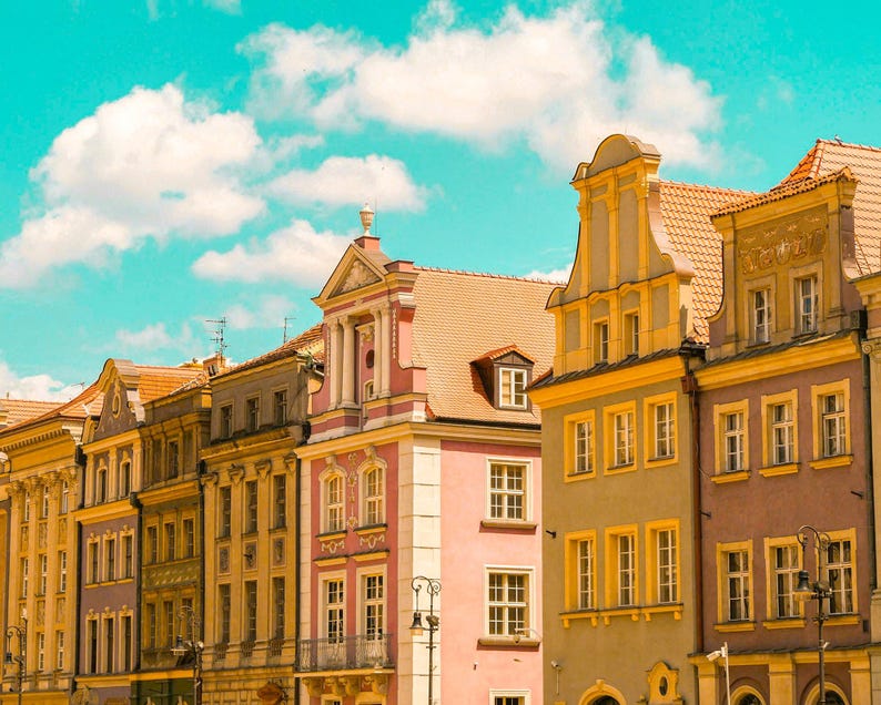 Pu&ograve; includere: Una fila di edifici a pi&ugrave; piani con facciate ornate nei toni del rosa, giallo e grigio, caratterizzati da tetti a capanna, finestre rettangolari con cornici gialle e dettagli architettonici decorativi sotto un cielo azzurro brillante.