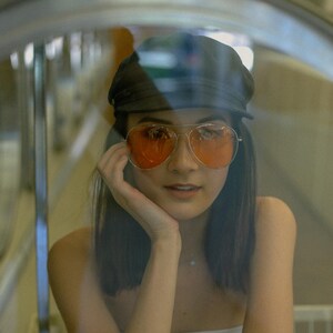 K&ouml;nnte beinhalten: Eine Frau mit dunklen Haaren, die eine schwarze M&uuml;tze und eine orange get&ouml;nte Sonnenbrille tr&auml;gt. Sie schaut in die Kamera und hat die Hand am Kinn.