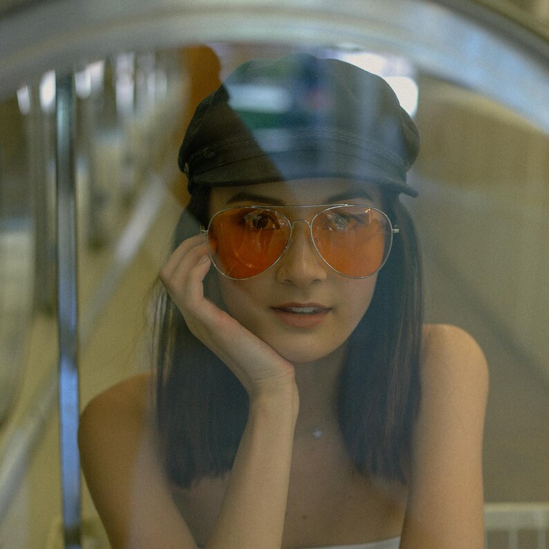 K&ouml;nnte beinhalten: Eine Frau mit dunklen Haaren, die eine schwarze M&uuml;tze und eine orange get&ouml;nte Sonnenbrille tr&auml;gt. Sie schaut in die Kamera und hat die Hand am Kinn.