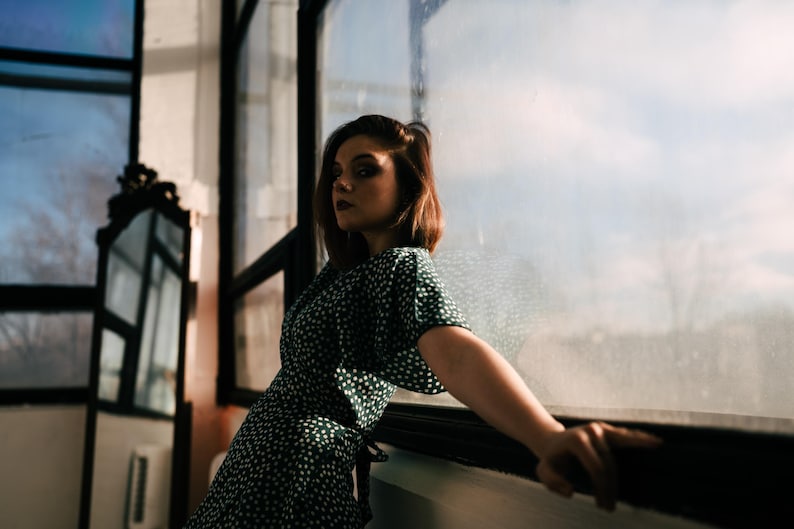 May include: A dark green dress featuring a white polka dot pattern, short sleeves, and a wrap-style front closure. The garment is constructed with a lightweight fabric and displays a consistent print throughout the bodice and sleeve sections.