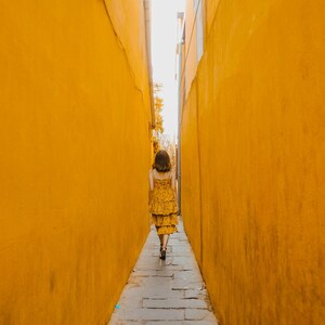 Pu&ograve; includere: Un abito midi a balze di colore giallo senape con un sottile motivo floreale scuro, caratterizzato da spalline sottili e un corpetto aderente, indossato mentre si cammina lungo uno stretto sentiero in pietra tra due alti muri gialli.