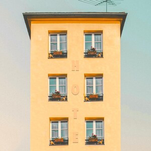 Pu&ograve; includere: Un'alta facciata di edificio gialla con sei finestre disposte in due colonne. Tra le colonne, le lettere H, O, T, E sono allineate verticalmente. Ogni finestra presenta una cornice scura e una fioriera marrone con piccoli fiori rossi.