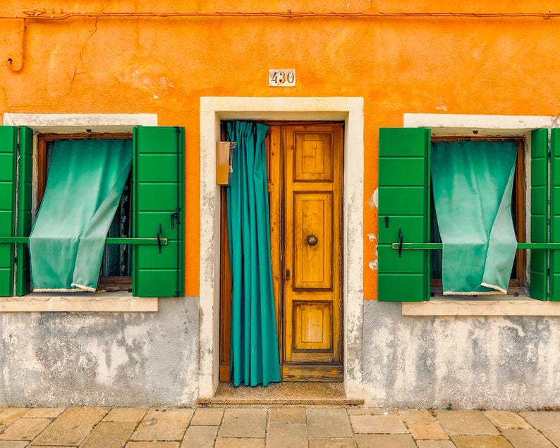 Pu&ograve; includere: Una facciata di edificio color arancione brillante con una porta centrale in legno dotata di una tenda color ottanio, affiancata da due finestre con persiane verdi e tende ottanio. Il numero 430 &egrave; visibile sopra la porta, su una base grigia.