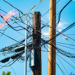Puede incluir: Un poste de madera desgastado marcado con el n&uacute;mero 657, con una caja de conexiones de metal gris y una densa red de cables el&eacute;ctricos negros que se cruzan contra un cielo azul claro, junto a un poste blanco liso y otro poste de madera.