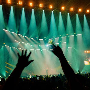 Pu&ograve; includere: Un palco da concerto illuminato da faretti color verde acqua e luci arancioni dall'alto. Il palco presenta un grande schermo centrale e display laterali che mostrano gli artisti. Sagome di spettatori con le mani alzate in primo piano.