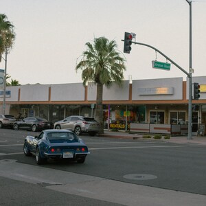 K&ouml;nnte beinhalten: Ein blauer klassischer Chevrolet Corvette Sportwagen, der auf einer Stra&szlig;e vor einem Geb&auml;ude mit einem Schild mit der Aufschrift "Vermietung" geparkt ist.