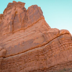 Puede incluir: Una imponente formaci&oacute;n rocosa de arenisca roja con estr&iacute;as verticales y texturas estratificadas, sobre un cielo azul claro. La enorme pared rocosa muestra patrones de erosi&oacute;n naturales, grietas profundas y un color rojo anaranjado c&aacute;lido.