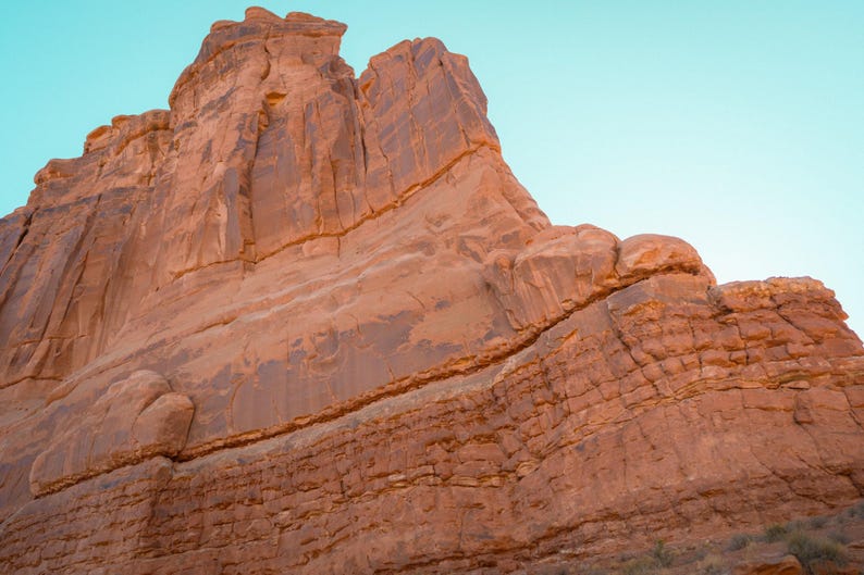 Puede incluir: Una imponente formaci&oacute;n rocosa de arenisca roja con estr&iacute;as verticales y texturas estratificadas, sobre un cielo azul claro. La enorme pared rocosa muestra patrones de erosi&oacute;n naturales, grietas profundas y un color rojo anaranjado c&aacute;lido.