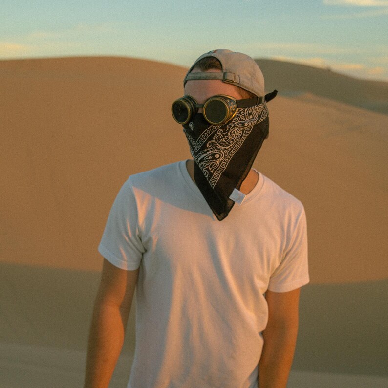 K&ouml;nnte beinhalten: Eine Person tr&auml;gt ein wei&szlig;es T-Shirt, ein schwarzes Bandana und eine Schutzbrille. Die Person steht in einer W&uuml;stenlandschaft.