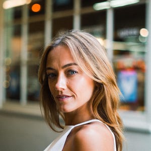 May include: A person with shoulder-length blonde hair and blue eyes wearing a white sleeveless top with a high neckline. The individual is positioned in profile, looking toward the camera with a neutral expression against a blurred background.