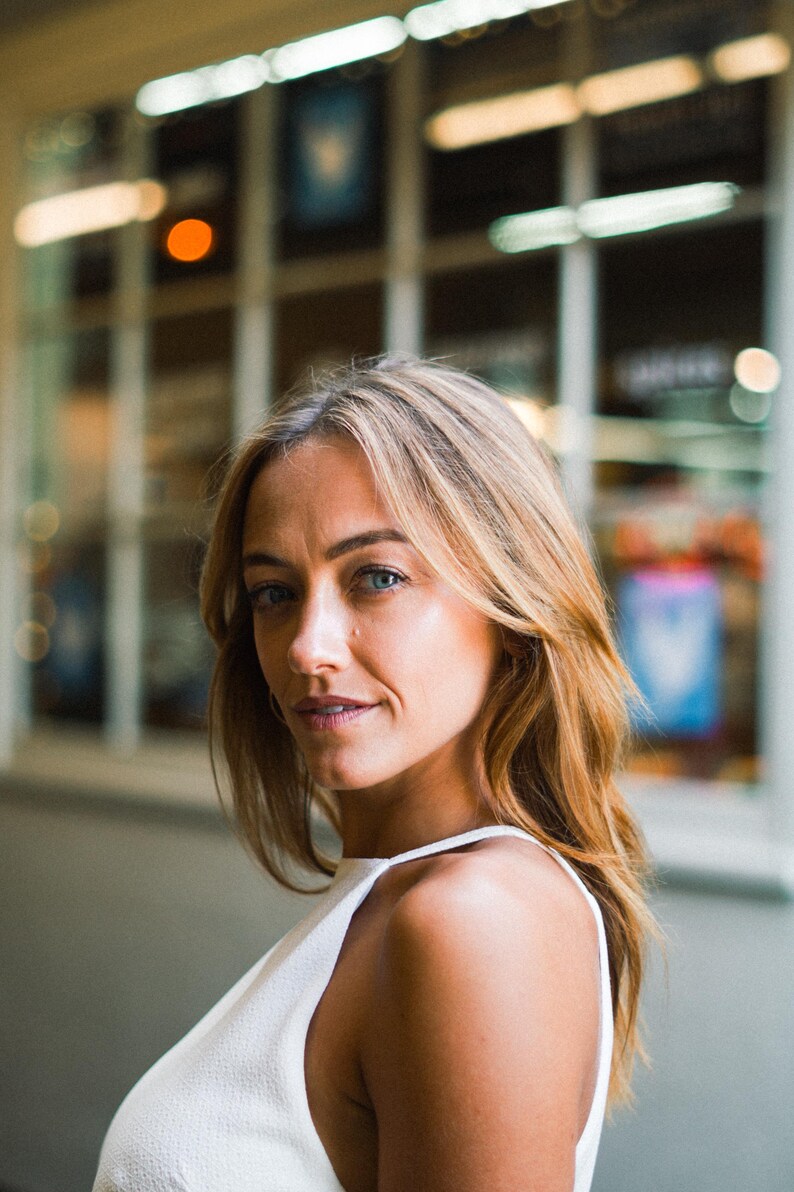 May include: A person with shoulder-length blonde hair and blue eyes wearing a white sleeveless top with a high neckline. The individual is positioned in profile, looking toward the camera with a neutral expression against a blurred background.