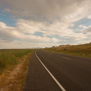 May include: A paved asphalt road stretches into the distance through a dry, grassy landscape under a cloudy sky. The road features a solid white line on the left edge and a broken yellow line in the center, surrounded by golden-brown vegetation.