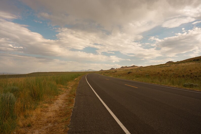 May include: A paved asphalt road stretches into the distance through a dry, grassy landscape under a cloudy sky. The road features a solid white line on the left edge and a broken yellow line in the center, surrounded by golden-brown vegetation.