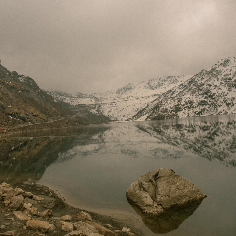 K&ouml;nnte beinhalten: Ein stiller See, der die schneebedeckten Berge in der Ferne spiegelt. Ein gro&szlig;er, grauer Felsen liegt im Vordergrund, teilweise im Wasser versunken.