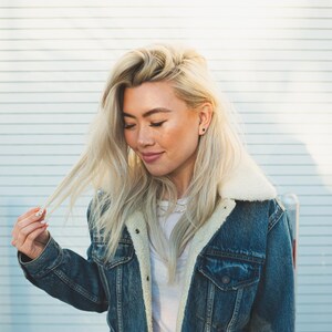 May include: A blue denim jacket featuring a white sherpa fleece collar and lining, metal button closures, and two chest pockets. The jacket is worn over a plain white t-shirt, showcasing a classic design with visible stitching and metal hardware.