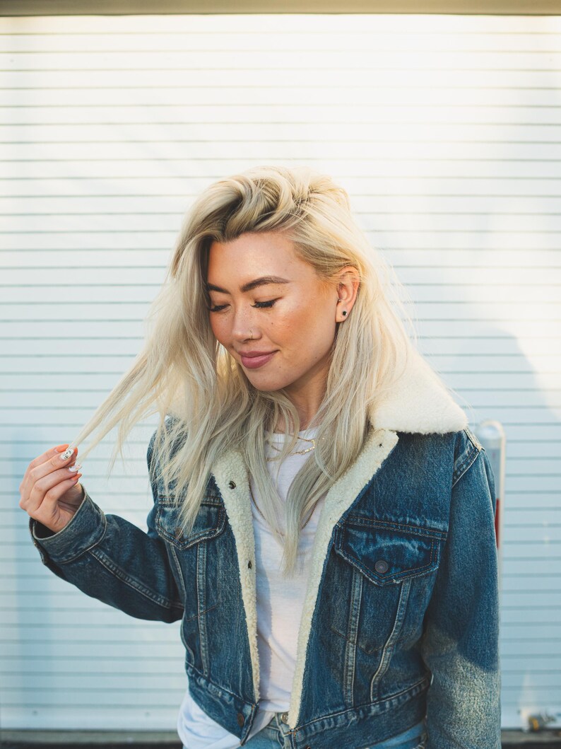 May include: A blue denim jacket featuring a white sherpa fleece collar and lining, metal button closures, and two chest pockets. The jacket is worn over a plain white t-shirt, showcasing a classic design with visible stitching and metal hardware.