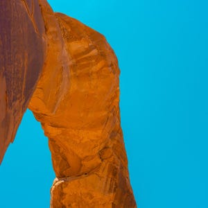 Pu&ograve; includere: Una formazione ad arco in arenaria naturale con una superficie ruvida color arancione-rosso. La struttura rocciosa presenta un'apertura curva contro un cielo azzurro limpido. La pietra mostra motivi geologici stratificati e ombre.