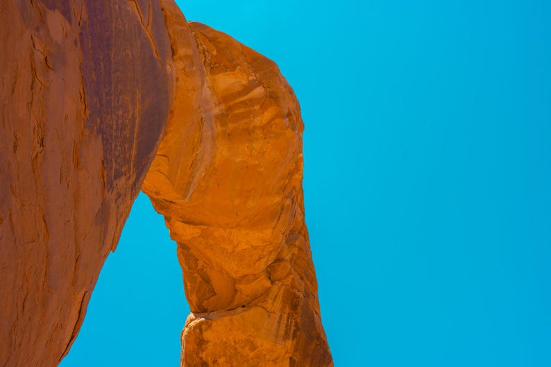 Pu&ograve; includere: Una formazione ad arco in arenaria naturale con una superficie ruvida color arancione-rosso. La struttura rocciosa presenta un'apertura curva contro un cielo azzurro limpido. La pietra mostra motivi geologici stratificati e ombre.