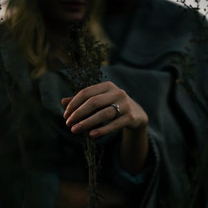 May include: A silver ring with a central stone and a twisted band design worn on a finger. The ring is positioned near dried, dark-colored plant stems, with a soft-focus grey fabric draped in the background under dim, natural lighting conditions.