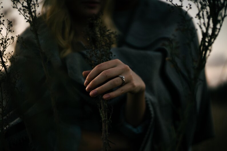 May include: A silver ring with a central stone and a twisted band design worn on a finger. The ring is positioned near dried, dark-colored plant stems, with a soft-focus grey fabric draped in the background under dim, natural lighting conditions.