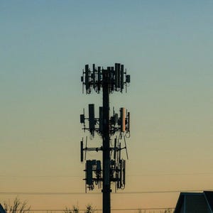 May include: A tall, dark gray cellular communication tower featuring multiple rectangular antennas and equipment panels mounted in three distinct tiers against a gradient sky transitioning from light blue to a warm orange sunset hue.