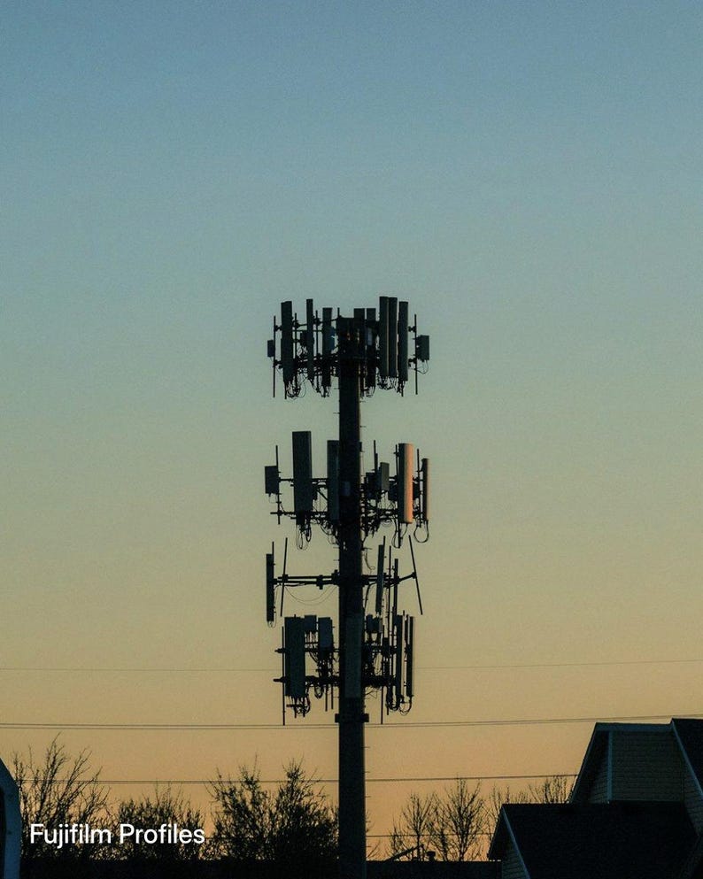 May include: A tall, dark gray cellular communication tower featuring multiple rectangular antennas and equipment panels mounted in three distinct tiers against a gradient sky transitioning from light blue to a warm orange sunset hue.