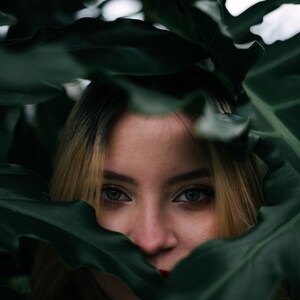 May include: A face framed by large, dark green, lobed tropical leaves. The eyes are visible, showing light irises and dark lashes, while the hair is blonde with darker roots. The foliage partially obscures the forehead and the lower face.