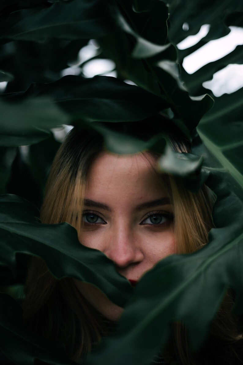May include: A face framed by large, dark green, lobed tropical leaves. The eyes are visible, showing light irises and dark lashes, while the hair is blonde with darker roots. The foliage partially obscures the forehead and the lower face.
