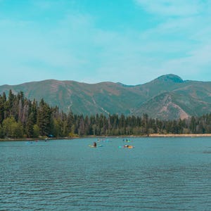 Pu&ograve; includere: Una vista panoramica di un lago calmo con diverse persone su tavole da stand-up paddle. La riva &egrave; fiancheggiata da una fitta foresta di alberi sempreverdi e grandi montagne ondulate si ergono sullo sfondo sotto un cielo azzurro.