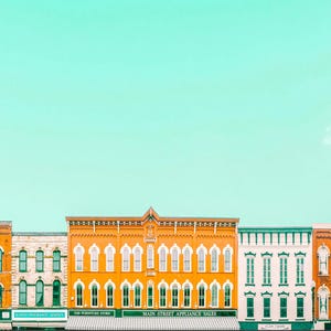 Pu&ograve; includere: Una fila di edifici storici in mattoni con finestre ad arco e vetrine sotto un cielo verde menta brillante. Gli edifici sono dipinti in tonalit&agrave; arancione, bianco e verde, con insegne di attivit&agrave; commerciali come Main Street Appliance Sales.