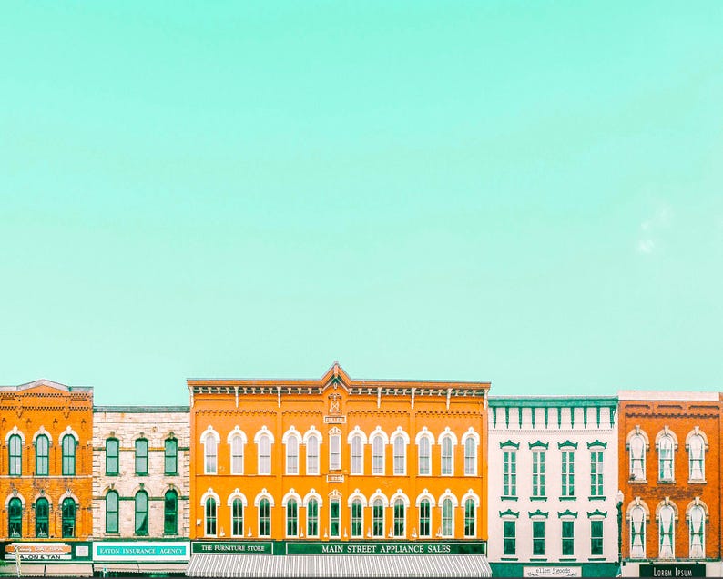 Pu&ograve; includere: Una fila di edifici storici in mattoni con finestre ad arco e vetrine sotto un cielo verde menta brillante. Gli edifici sono dipinti in tonalit&agrave; arancione, bianco e verde, con insegne di attivit&agrave; commerciali come Main Street Appliance Sales.