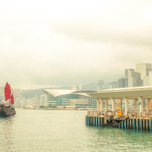 Puede incluir: Un junco chino tradicional con velas de color rojo brillante navegando por las aguas del puerto Victoria en Hong Kong. Al fondo, se aprecian el moderno Centro de Convenciones y Exposiciones de Hong Kong y un denso horizonte de rascacielos.