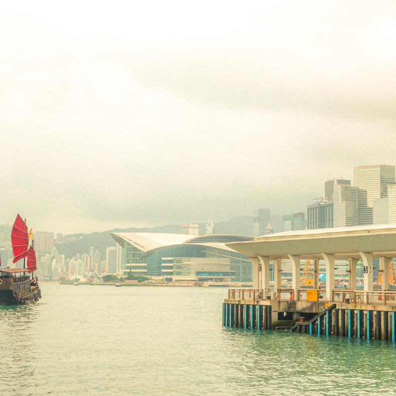 Puede incluir: Un junco chino tradicional con velas de color rojo brillante navegando por las aguas del puerto Victoria en Hong Kong. Al fondo, se aprecian el moderno Centro de Convenciones y Exposiciones de Hong Kong y un denso horizonte de rascacielos.