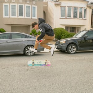 K&ouml;nnte beinhalten: Eine Person in einem schwarzen Hoodie und braunen Hosen springt &uuml;ber ein Skateboard mit regenbogenfarbenem Griptape. Das Skateboard befindet sich auf einer grauen Asphaltstra&szlig;e vor einem Auto.