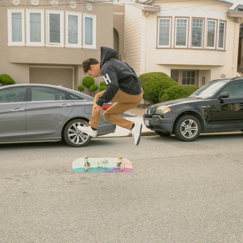 K&ouml;nnte beinhalten: Eine Person in einem schwarzen Hoodie und braunen Hosen springt &uuml;ber ein Skateboard mit regenbogenfarbenem Griptape. Das Skateboard befindet sich auf einer grauen Asphaltstra&szlig;e vor einem Auto.