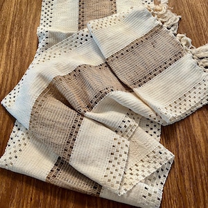May include: Cream and brown woven table runner with a rectangular design. The runner features alternating stripes of cream-colored open weave and brown woven sections. The edges are finished with a fringe.
