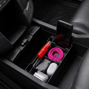 May include: A black plastic center console organizer tray installed in a vehicle, featuring multiple compartments holding a smartphone, a bright pink coiled charging cable, a red spray bottle, a white earbud case, and other small personal items.