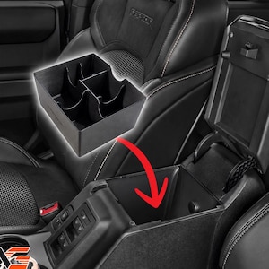 May include: A black plastic center console organizer tray with multiple compartments designed to fit into the deep storage bin of a vehicle. The tray is shown hovering above the open console lid, with a red arrow indicating placement.
