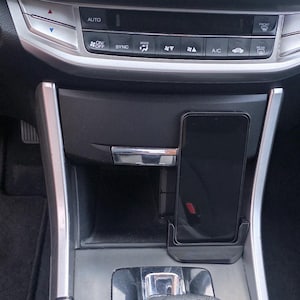 Peut inclure: Un smartphone noir est monté dans un support de téléphone sur le tableau de bord d'une voiture. L'intérieur de la voiture présente une palette de couleurs noire et argentée, avec divers boutons et commandes visibles. Le levier de vitesses est au premier plan.